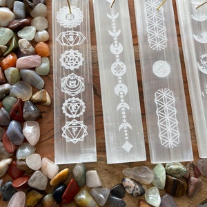 May include: Four white selenite incense holders with different etched designs. The designs include the seven chakras, moon phases, the flower of life, and a geometric pattern. The holders are on a wooden surface with colorful stones scattered around them.