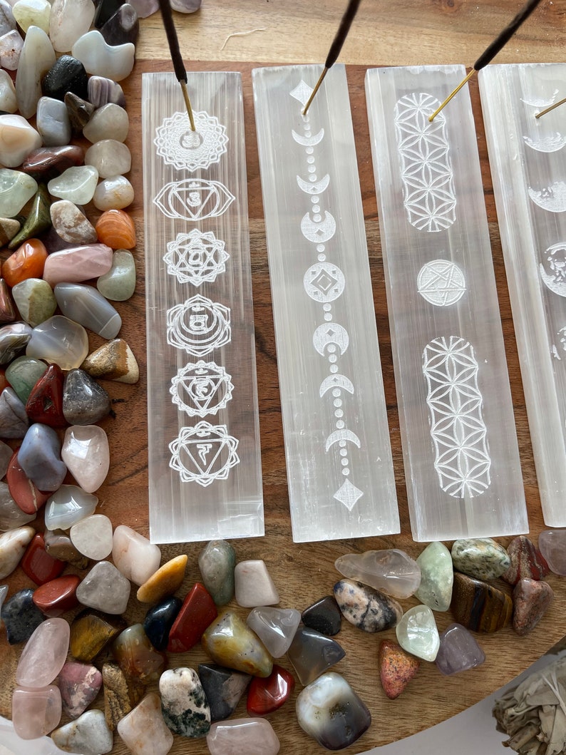 May include: Four white selenite incense holders with different etched designs. The designs include the seven chakras, moon phases, the flower of life, and a geometric pattern. The holders are on a wooden surface with colorful stones scattered around them.