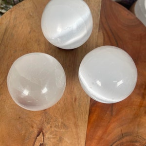 Op de afbeelding: Drie witte seleniet bollen op een houten oppervlak. De bollen zijn glad en hebben een doorschijnende uitstraling.