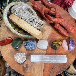 May include: A wooden tray with a variety of crystals, including amethyst, tiger's eye, and selenite. There is also a bundle of sage and a wooden wand.