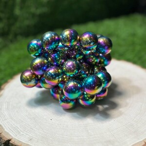 K&ouml;nnte beinhalten: Eine w&uuml;rfelf&ouml;rmige Skulptur aus zahlreichen irisierenden, kugelf&ouml;rmigen Magnetkugeln. Die Kugeln zeigen ein lebendiges Farbspektrum, darunter Blau-, Gr&uuml;n-, Lila- und Gelbt&ouml;ne. Die Skulptur ruht auf einer nat&uuml;rlichen Holzscheibe mit verschwommenem gr&uuml;nem Hintergrund.