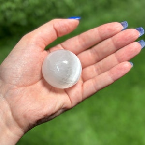 Op de afbeelding: Een witte selenietbol, een gladde, gepolijste kristallen bol, vastgehouden in een hand tegen een groene achtergrond.