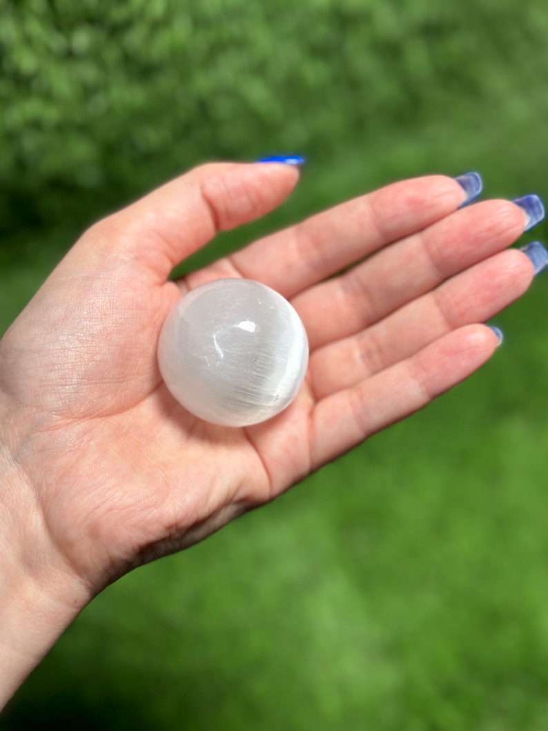 Op de afbeelding: Een witte selenietbol, een gladde, gepolijste kristallen bol, vastgehouden in een hand tegen een groene achtergrond.