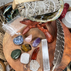 May include: A collection of crystals and stones, including amethyst, rose quartz, selenite, and tiger's eye, arranged on a wooden surface with a sage smudge stick and a feather.