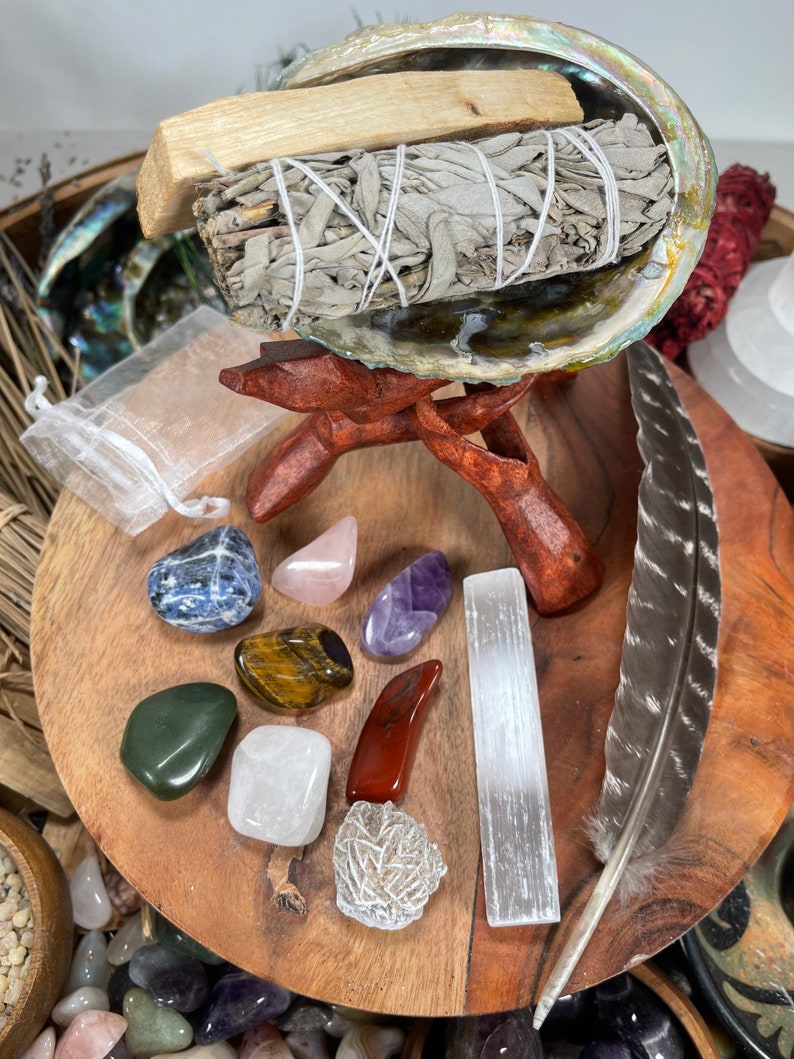 May include: A collection of crystals and stones, including amethyst, rose quartz, selenite, and tiger's eye, arranged on a wooden surface with a sage smudge stick and a feather.