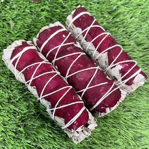 May include: Three white sage smudge sticks wrapped in dried rose petals. The sticks are tied with white string and are arranged in a row on a green surface.