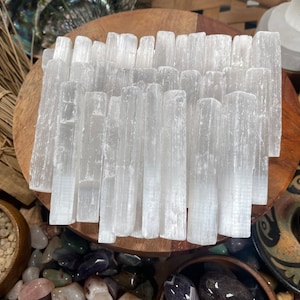 May include: A collection of white selenite wands, also known as selenite sticks, arranged on a wooden surface. The wands are smooth and have a natural, striated texture.