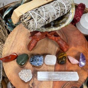 May include: A wooden stand with a seashell bowl holding a bundle of white sage and a wooden stick. The stand is surrounded by various crystals, including red, green, blue, white, brown, pink, and purple.