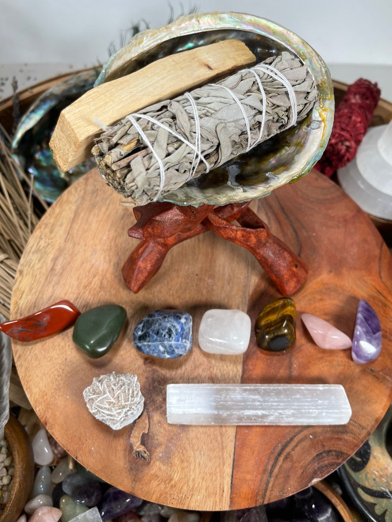 May include: A wooden stand with a seashell bowl holding a bundle of white sage and a wooden stick. The stand is surrounded by various crystals, including red, green, blue, white, brown, pink, and purple.
