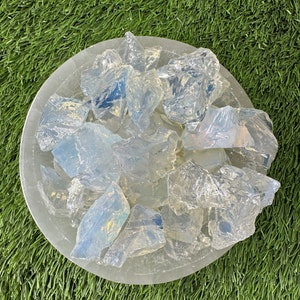 May include: A bowl filled with clear, iridescent opalite chips. The bowl is white and has a smooth, rounded surface. The opalite chips are various sizes and shapes, and they shimmer with a rainbow of colors.