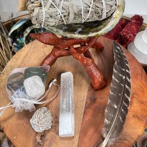 May include: A wooden stand with a white sage smudge stick in a seashell, a clear quartz crystal, a small white stone, and a brown feather.