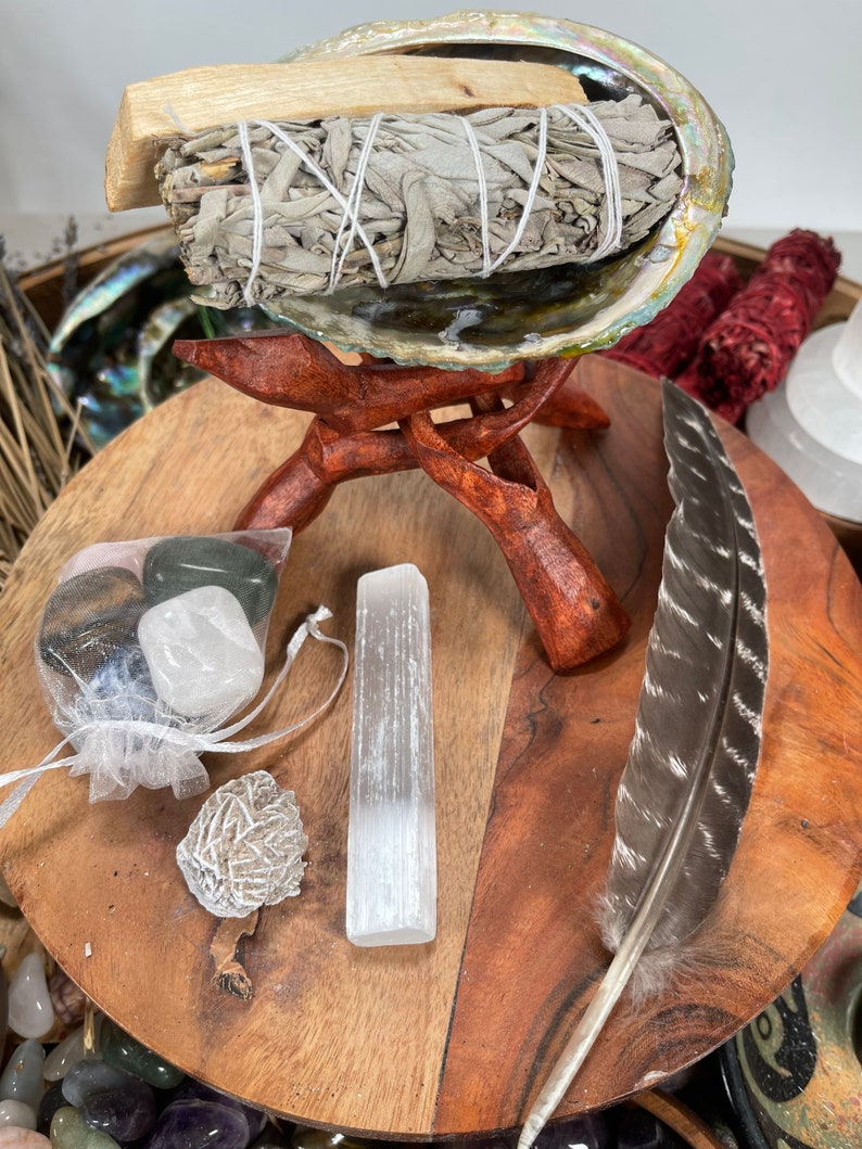 May include: A wooden stand with a white sage smudge stick in a seashell, a clear quartz crystal, a small white stone, and a brown feather.