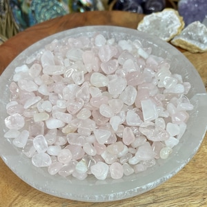 May include: A white bowl filled with small, light pink rose quartz chips. The bowl is sitting on a wooden surface.