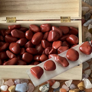 May include: A wooden box filled with red jasper tumbled stones. The box is open and the stones are visible. There is a white crystal wand with red jasper stones on top of it.
