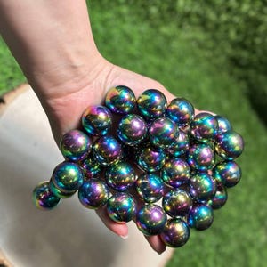 K&ouml;nnte beinhalten: Eine Ansammlung schillernder, kugelf&ouml;rmiger Perlen in Lila, Gr&uuml;n und Blau. Die Perlen werden in einer Hand gehalten, mit einem verschwommenen gr&uuml;nen Hintergrund. Die Perlen haben einen metallischen Glanz.