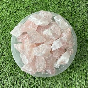 May include: A bowl filled with rose quartz crystals. The crystals are a light pink color and have a natural, rough texture. The bowl is sitting on a green surface.