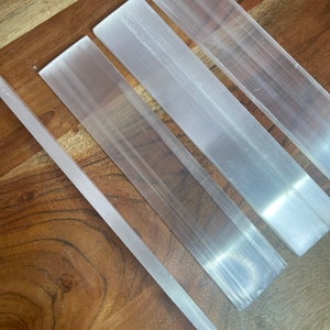 May include: Four selenite wands of varying lengths, arranged on a wooden surface. The wands are smooth and white with a translucent appearance.