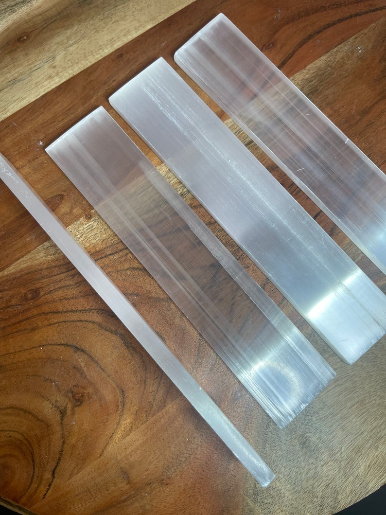 May include: Four selenite wands of varying lengths, arranged on a wooden surface. The wands are smooth and white with a translucent appearance.