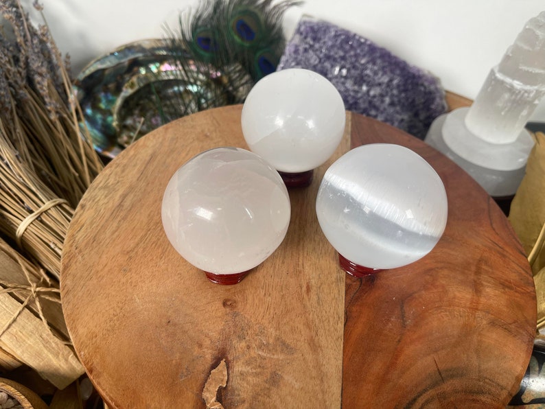 Op de afbeelding: Drie witte seleniet bollen op houten standaards. De bollen zijn glad en gepolijst, met een lichte glans. De standaards zijn donker roodbruin.