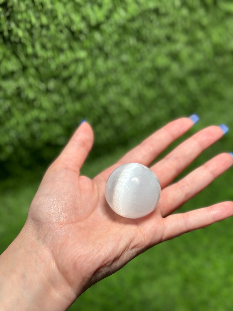 Op de afbeelding: Een witte selenietbol, ook wel bekend als maansteen, wordt in een hand gehouden. De bol is glad en heeft een doorschijnende uitstraling.