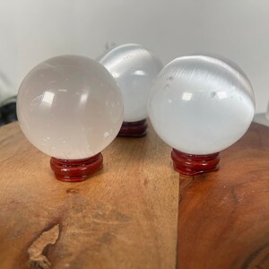 Op de afbeelding: Drie witte selenietkristallen bollen op houten standaards. De bollen zijn glad en doorschijnend, met een lichte glans.