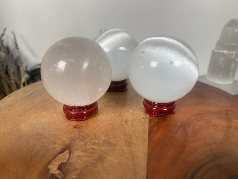 Op de afbeelding: Drie witte selenietkristallen bollen op houten standaards. De bollen zijn glad en doorschijnend, met een lichte glans.