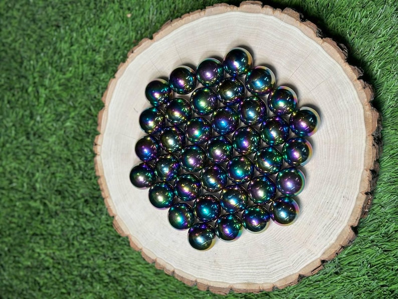 K&ouml;nnte beinhalten: Eine Ansammlung schillernder Murmeln, die eine Vielzahl von Farben reflektieren, auf einer Holzscheibe angeordnet. Die Murmeln, mit Farbt&ouml;nen von Blau, Gr&uuml;n und Lila, sind vor einem Hintergrund aus gr&uuml;nem Gras platziert und erzeugen eine lebendige Darstellung.