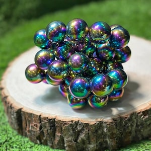K&ouml;nnte beinhalten: Eine Ansammlung schillernder Magnetkugeln, die ein Farbspektrum von Blau &uuml;ber Gr&uuml;n bis Violett zeigen. Die Kugeln sind kompakt angeordnet und liegen auf einer nat&uuml;rlichen Holzscheibe. Der Hintergrund ist gr&uuml;n.