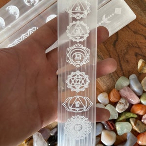 May include: White selenite wand with seven chakra symbols etched into the surface. The wand is resting on a bed of colorful tumbled stones.