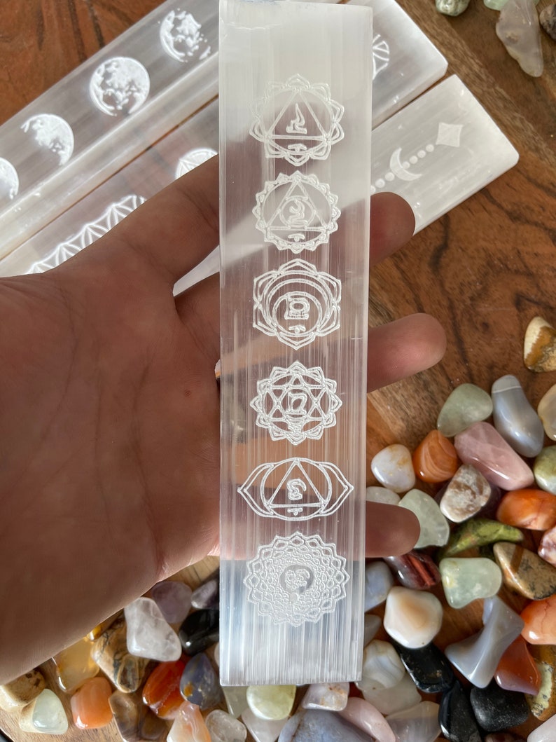 May include: White selenite wand with seven chakra symbols etched into the surface. The wand is resting on a bed of colorful tumbled stones.