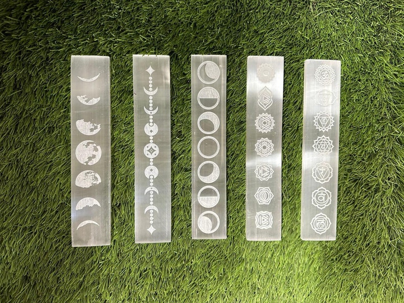 May include: Five white selenite wands with different etched designs. The designs include phases of the moon, a celestial chain, a full moon, chakra symbols, and a set of seven chakras.