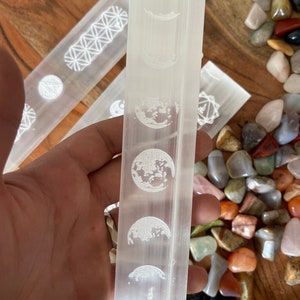 May include: White selenite wand with a moon phase design etched into the surface. The wand is approximately 6 inches long and is surrounded by a variety of colorful stones.