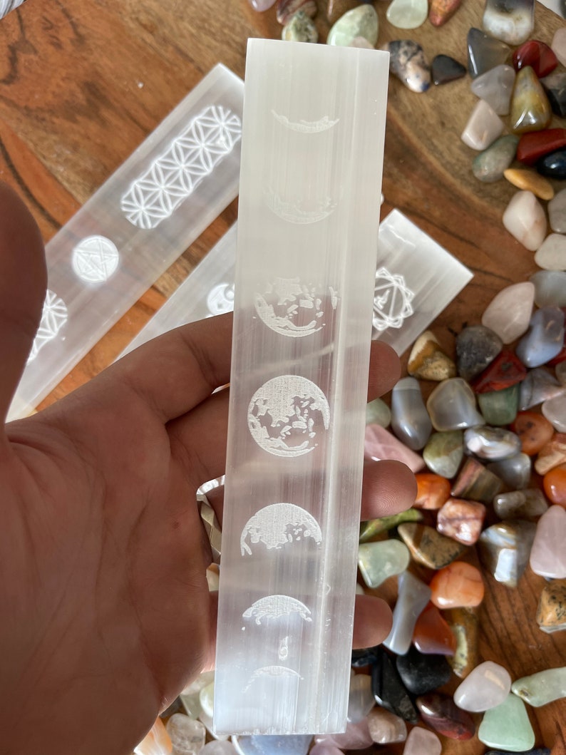 May include: White selenite wand with a moon phase design etched into the surface. The wand is approximately 6 inches long and is surrounded by a variety of colorful stones.