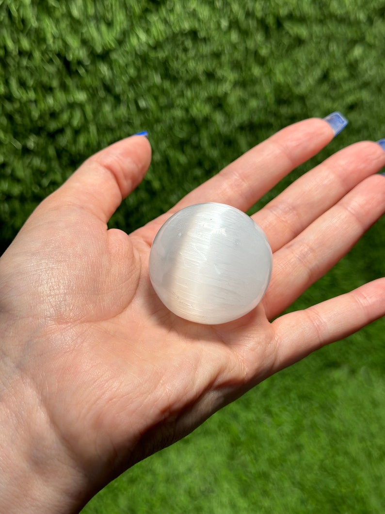 Op de afbeelding: Een witte selenietbol, ook wel een selenietkogel genoemd, wordt in een hand gehouden. De bol is glad en heeft een gestreepte textuur.