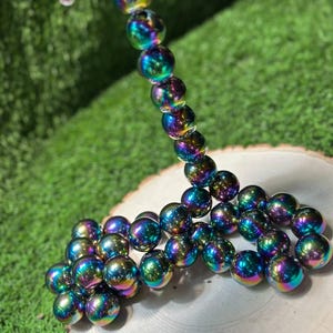 K&ouml;nnte beinhalten: Eine Halskette oder ein Armband mit irisierenden, kugelf&ouml;rmigen Perlen in Blau-, Gr&uuml;n- und Lilat&ouml;nen. Die Perlen, mit einem Durchmesser von ca. 2 cm, sind aufgereiht, ein Teil wird von einer Hand gehalten. Sie haben ein reflektierendes, metallisches Aussehen.