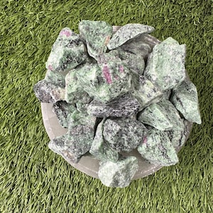 May include: A collection of green and black rough zultanite gemstones in a white bowl. The stones have a speckled appearance with hints of pink.