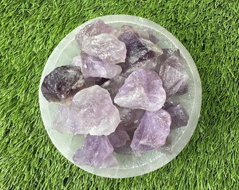 May include: A white bowl filled with amethyst crystals. The crystals are various shades of purple and have a rough, natural texture. The bowl is sitting on a green grassy surface.