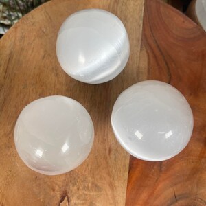 Op de afbeelding: Drie witte seleniet bollen op een houten oppervlak. De bollen zijn glad en hebben een licht iriserende glans.