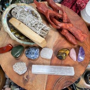 May include: A wooden tray with a variety of crystals, including amethyst, tiger's eye, rose quartz, and selenite. There is also a bundle of sage and a wooden stand.