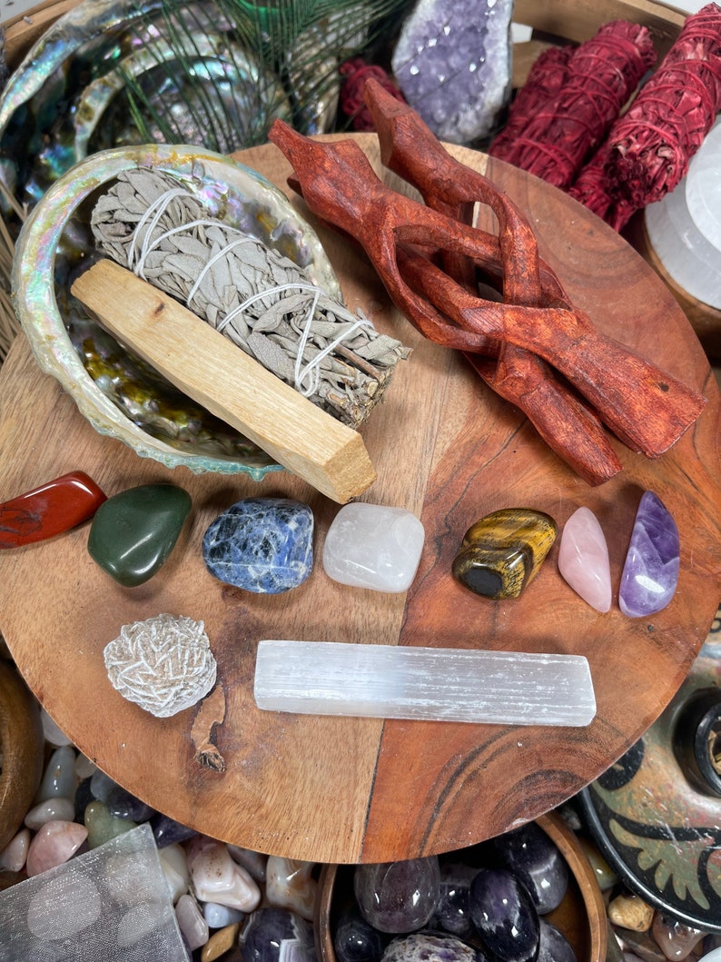 May include: A wooden tray with a variety of crystals, including amethyst, tiger's eye, rose quartz, and selenite. There is also a bundle of sage and a wooden stand.