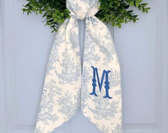 May include: A white and blue toile fabric bow with a blue embroidered letter "M" in the center. The bow is tied around a green wreath.