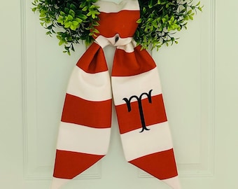 May include: Red and white striped fabric bow with a black embroidered letter "T" in the center. The bow is tied around a green wreath.