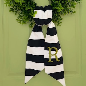 May include: A black and white striped fabric bow with the letter "R" embroidered in green. The bow is hanging on a green door.