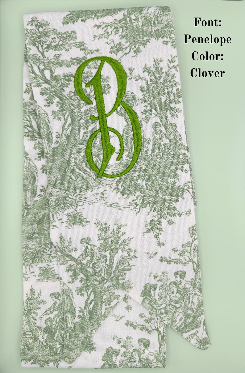 May include: A white fabric with a green toile pattern and a green embroidered letter "B" in the Penelope font. The text "Font: Penelope Color: Clover" is visible in the upper right corner of the image.