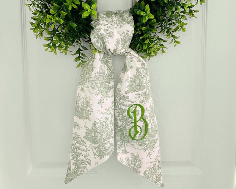 May include: A white and green toile fabric bow with a green embroidered letter "B" on the right side. The bow is tied around a green wreath.