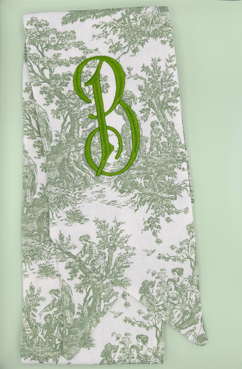 May include: A white fabric tea towel with a green toile pattern and a green embroidered letter "B".