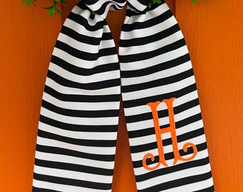 May include: A fabric ribbon featuring a black and white horizontal striped pattern. The ribbon is tied in a knot at the top and displays a bright orange embroidered monogram letter H on the lower right tail of the fabric.