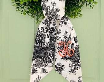 May include: A white fabric ribbon with a black and white toile pattern. The ribbon is tied into a bow and has a large orange embroidered letter "H" in the center.