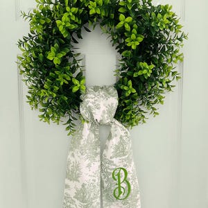 May include: A green leafy wreath with a white and green toile fabric bow with a green embroidered letter "B" on it.