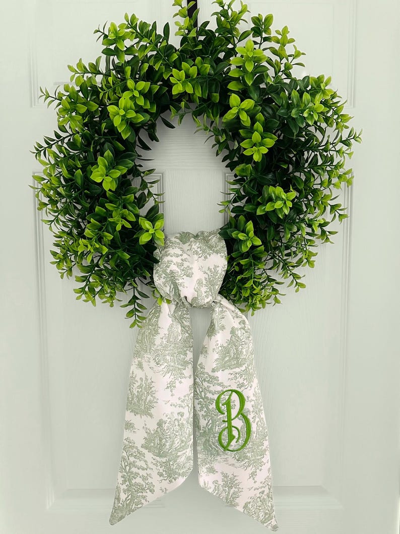 May include: A green leafy wreath with a white and green toile fabric bow with a green embroidered letter "B" on it.